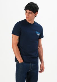 Homme aux cheveux foncés portant un t-shirt bleu marine de Tommy Jeans et un jean sombre, une main dans la poche, regardant sur le côté sur fond blanc.
