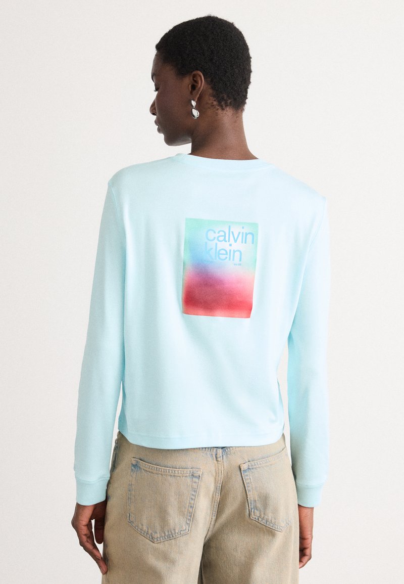 Hellblaues Langarm-Sweatshirt mit einem rechteckigen, mehrfarbigen Motiv mit der Aufschrift "Calvin Klein" auf dem Rücken. Weicher Stoff und lockere Passform.