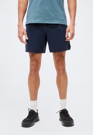 Mand iført marineblå shorts, petrolfarvet t-shirt, hvide sokker og sorte sneakers, som står mod en ensartet lys baggrund.