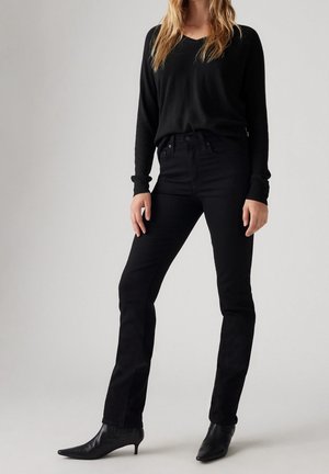 Vrouw die een zwart topje met lange mouwen draagt, hoge taille zwarte skinny jeans en zwarte puntige enkellaarsjes, staand tegen een effen achtergrond.