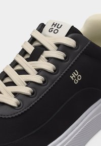 Μαύρο αθλητικό παπούτσι με εκρού κορδόνια, λευκή σόλα και λογότυπο "HUGO" στη γλώσσα και στο πλάι, σε κοντινή λήψη της περιοχής των κορδονιών.