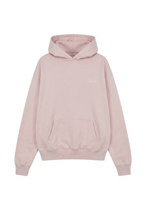 Ung mand iført en lys pink hættetrøje, grå løstsiddende bukser og beige sneakers, står med hænderne i lommerne mod en hvid baggrund.