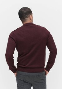 Burgundy neulepaita, jossa on polo-kaulus, ribattu hihansuut ja helma, päällä harmaat housut, kuvattu takaa yksinkertaista taustaa vasten.