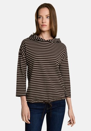 Woman wearing a brown and white horizontal striped long-sleeve top with a hood and dark blue jeans, standing against a light background.