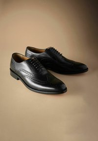 Zwarte lederen brogue nette schoenen met decoratieve perforaties en veters, weergegeven op een neutrale beige ondergrond.