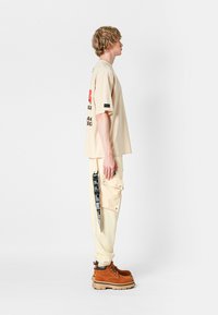 Camiseta beige de gran tamaño con estampado gráfico, combinada con pantalones cargo crema que tienen acentos de texto negro y un bolsillo lateral. Botas marrones.