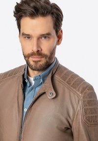 Giacca di pelle marrone con chiusura a zip, colletto a scatto e dettagli testurizzati sulle spalle, indossata sopra una camicia blu con bottoni.