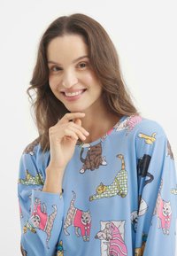 Niebieska piżama z luźnym krojem, ozdobiona zabawnym wzorem kotów w różnych pozach. Materiał wydaje się miękki, z okrągłym dekoltem i długimi rękawami.