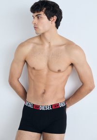Boxers noirs avec une ceinture de marque rouge et blanche. Le modèle a une silhouette tonique, posé de profil sur un fond clair.