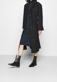 Cappotto a righe nere con un design sartoriale, bottoni dorati, indossato sopra un vestito aderente blu navy. Stivaletti neri lucidi alla caviglia completano l'outfit.