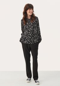 Zwarte blouse met lange mouwen en witte stippen, met een ronde hals. Gepaarde met zwarte broek en zwart-witte sneakers.