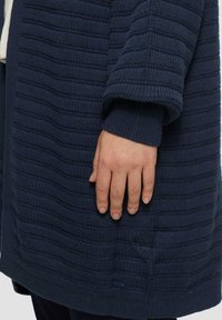 Cardigan en tricot bleu marine avec des rayures horizontales texturées, doté de poignets côtelés. Une main repose sur le tissu, mettant en avant sa douceur.