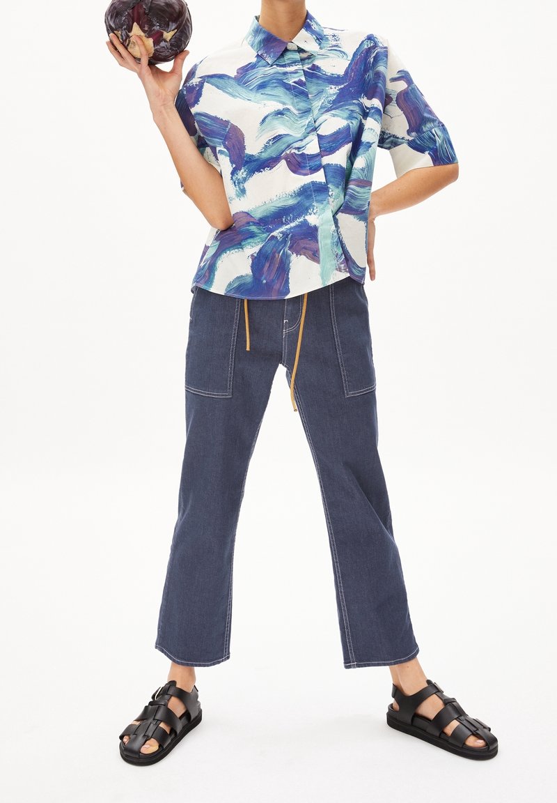 Personne portant une chemise à motif abstrait bleu et blanc, un pantalon large foncé et des sandales noires, tenant un chou violet d'une main.