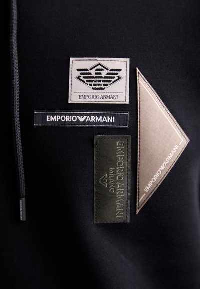 Svart tyg med olika Emporio Armani-märken: en vit logopatch, mörka och ljusa rektangulära etiketter och en beige rombformad detalj.