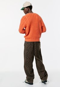 Orange kabelstickad tröja med ribbad fåll, matchad med mönstrade bruna baggybyxor och krämfärgade skor. Modellen bär en beige keps.