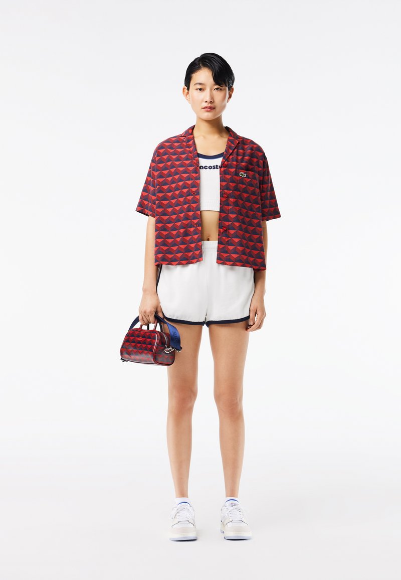 Rot und marineblau gemustertes Kurzarmhemd, weißes kurzes Oberteil, weiße Shorts mit marineblauen Akzenten, kombiniert mit einer kleinen roten Handtasche.