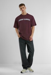 Burgundijas T-krekls ar "URBAN CLASSICS" logotipu gaiši zilā krāsā, saskaņots ar tumšiem džinsiem un pelēki zaļām sporta apavām. Ikdienišķs, brīvs piegriezums.