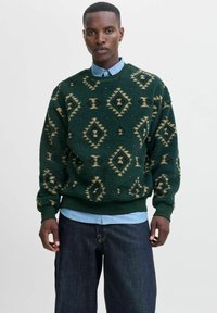 Pull en polaire vert avec des motifs en diamant et en zigzag beige, poignets et ourlet côtelés, superposé à une chemise à col bleu clair, associé à un jean foncé.