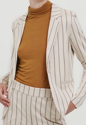 Vrouw draagt een crèmekleurig pak met dunne bruine verticale strepen en een mosterdkleurige coltrui, met één hand in de zak en de andere die het jasje rechtzet.