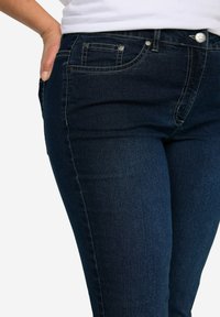 Tmavomodré džínsy s úzkym strihom, s tradičným päť vreckovým dizajnom, kontrastným prešívaním a kovovými detailmi na gombíkoch.