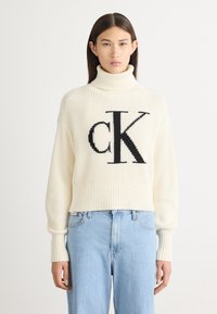 Cremfarbener gerippter Rollkragenpullover mit schwarzem "CK"-Logo auf der Vorderseite, kombiniert mit hellblauen Jeans. Weicher Stoff, lockere Passform.