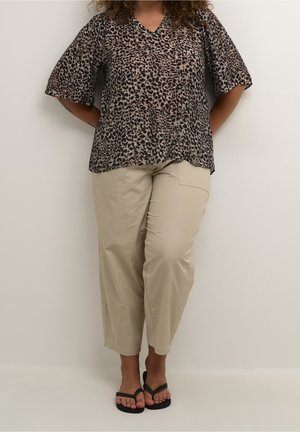 Person iført en bluse med leopardprint med albueærmer, beige korte bukser og sorte flip-flops, stående mod en ensfarvet væg.