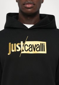 Felpa nera con un ampio logo dorato "just/cavalli". Il tessuto sembra morbido, con una vestibilità rilassata e un cappuccio con cordino.