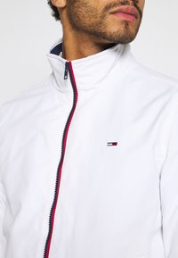 Chaqueta blanca con cremallera y cuello alto, detalle en cremallera roja y azul marino, y un pequeño logo bordado en el área del pecho izquierdo. Tejido ligero.
