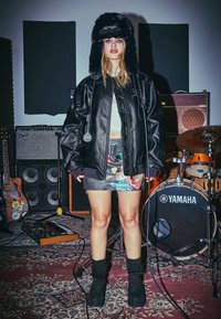 Černá kožená oversized bunda, vzorovaná mini sukně s barevnými motivy, huňatá černá čepice, černé boty, mikrofon v ruce, pozadí hudebního studia.