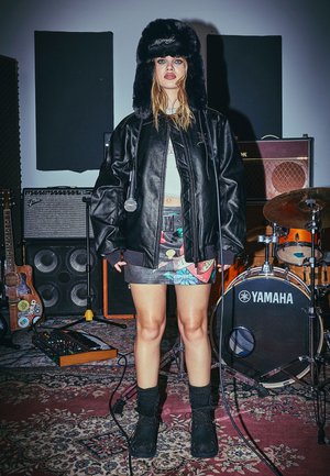 Chaqueta de cuero negra oversize, falda corta patterned con diseños coloridos, sombrero negro de piel, botas negras, micrófono en mano, fondo de estudio de música.