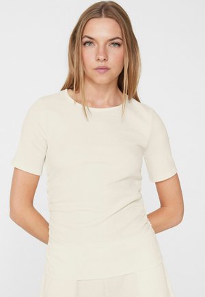 Jeune femme aux longs cheveux blonds portant un haut texturé à manches courtes, blanc cassé, sur un fond clair uni.
