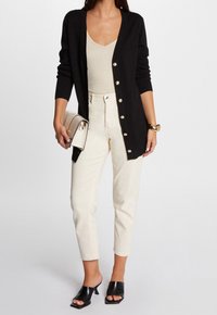 Un cardigan noir avec des boutons dorés, un haut crème, un pantalon beige taille haute et des mules noires ouvertes, portant un sac à main rayé beige.
