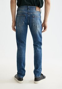 Vaqueros de mezclilla azul con un diseño ajustado, con bolsillos traseros y una etiqueta de cuero en la cintura. Llevados con zapatillas negras.