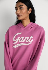 Pink hooded sweatshirt featuring a large white "Gant" logo and a smaller text "1949 NH. CT." on the front. Soft material with a relaxed fit.