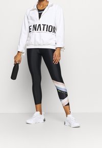 Biała bluza z zamkiem błyskawicznym z czarnym napisem, czarne legginsy z kolorowymi diamentowymi wzorami i białe sportowe buty; trzymająca czarną butelkę na wodę.