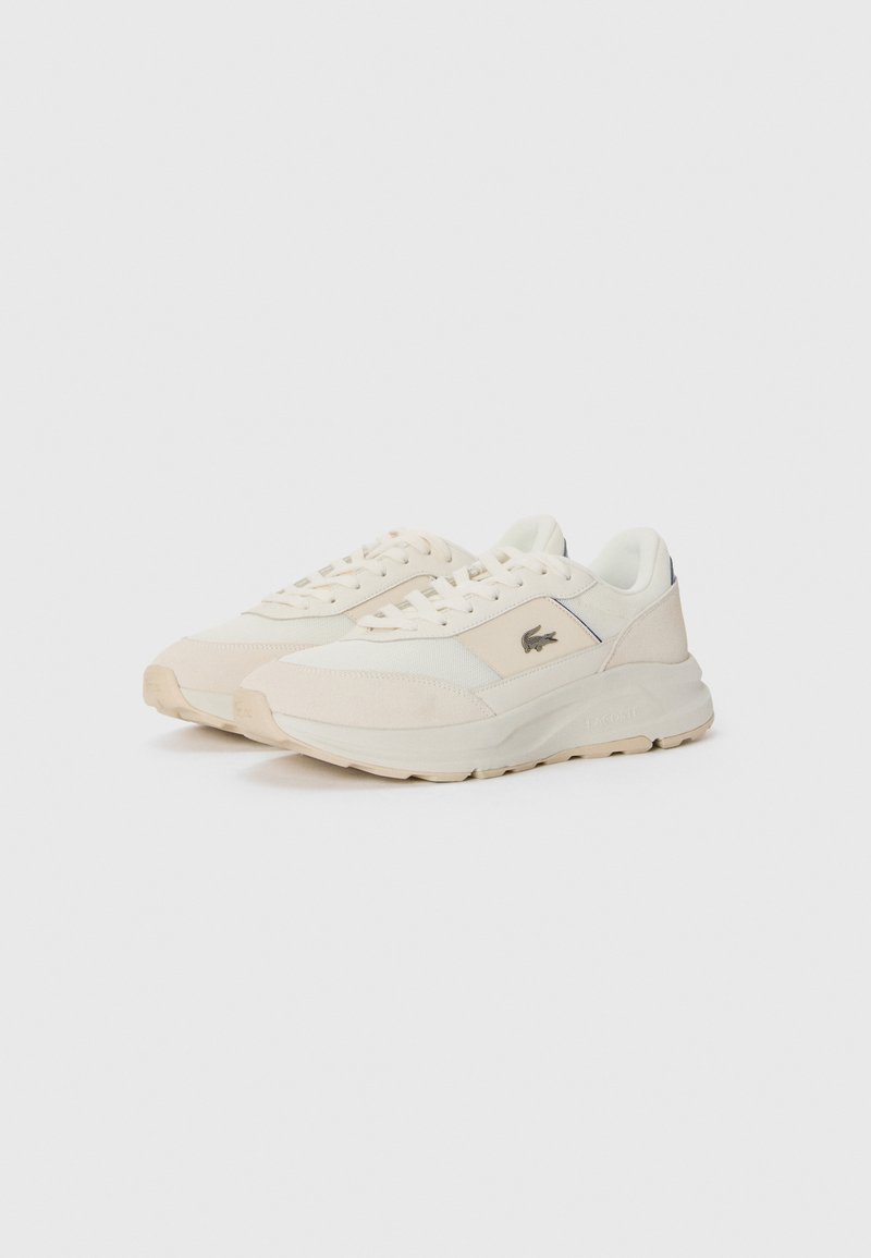 Une paire de baskets Lacoste de couleur crème avec des accents en suède beige, des panneaux en mesh et un logo sur le côté, présentée sur un fond blanc.