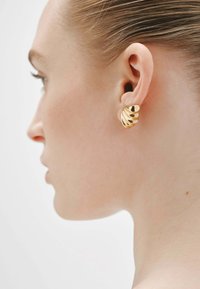 Pendientes en tono dorado con un diseño similar a una concha, con surcos curvados y recortes, exhibidos en la oreja de una modelo. Acabado de metal suave.