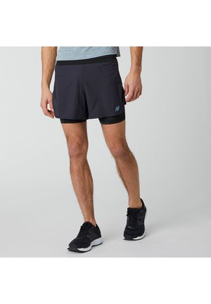 Mężczyzna w czarnych dwuwarstwowych szortach do biegania i czarnych butach do biegania stoi na jednolitym jasnoszarym tle.