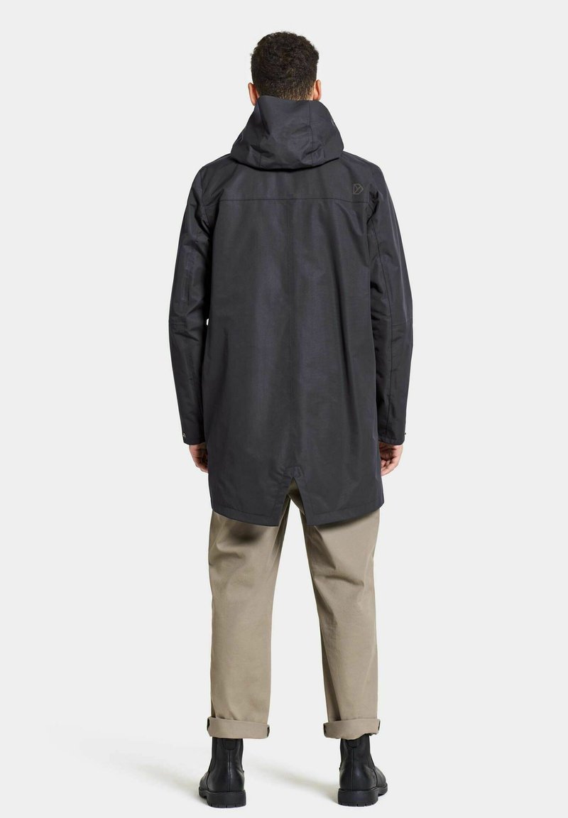 Schwarze wasserdichte Jacke mit Kapuze, lockerer Passform, längerem Rücken und seitlichem Schlitz, kombiniert mit beigen Hosen und schwarzen Stiefeln.