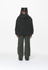 Hoodie in pile verde oversize con tasca a marsupio e cappuccio regolabile con cordino, abbinato a pantaloni olive verdi ampi. Superficie texture, vestibilità casual.
