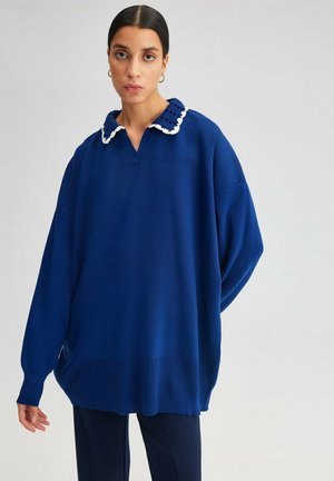 Femme aux cheveux foncés portant un pull bleu oversize avec un col tricoté blanc et bleu, debout devant un fond clair uni.