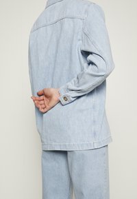 Jasnoniebieska kurtka jeansowa z długimi rękawami, mankietem zapinanym na guziki i detalem na tyle. Do kompletu spodnie w tym samym jasnoniebieskim kolorze.