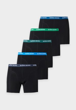 Homme athlétique debout, pieds nus, portant un boxer noir avec une taille verte sur un fond gris clair uni.
