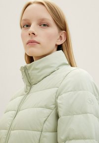 Helle grüne, gepolsterte Jacke mit hohem Kragen, ausgestattet mit einem Reißverschluss vorne und einem Logo-Akzent am Ärmel. Glatte, glänzende Textur.