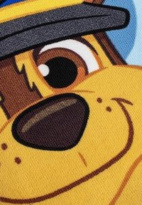 Personnage de chien cartoon, avec un grand visage souriant, une fourrure brun et jaune, des yeux noirs et un chapeau gris sur fond bleu.