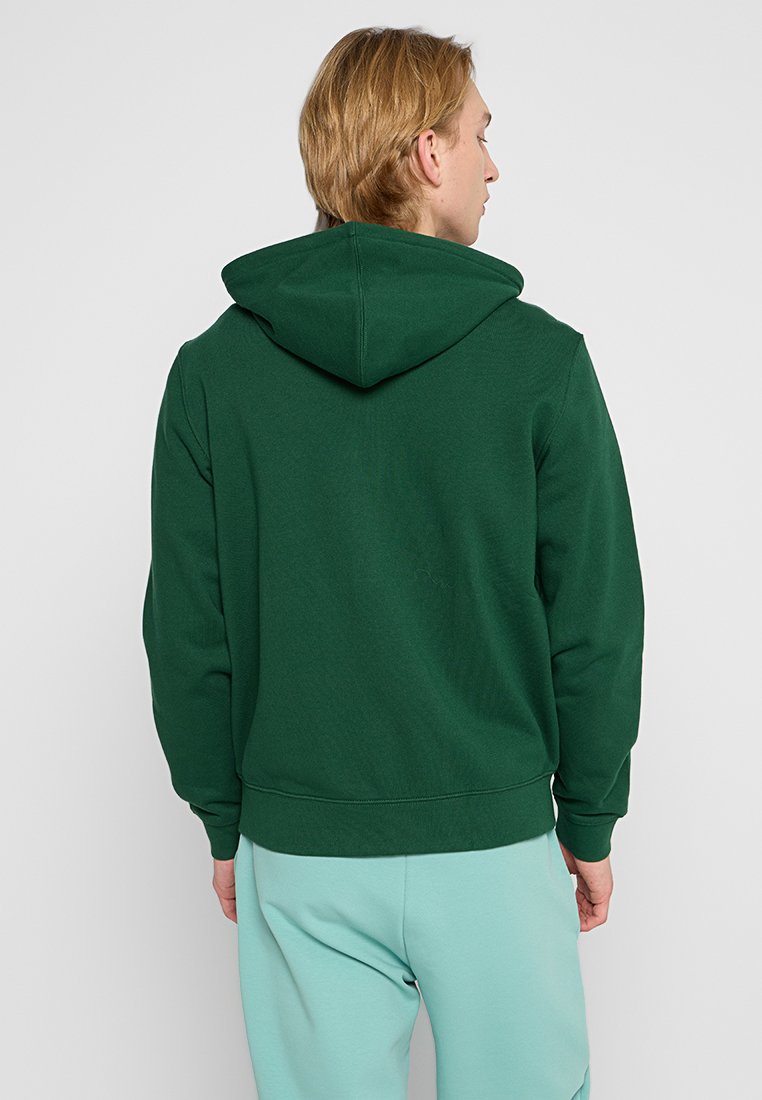 Sweat à capuche vert avec cordon de serrage, poignets et ourlet côtelés, présentant une texture lisse. Porté avec un pantalon de jogging bleu clair.