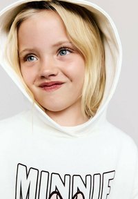 Haut à capuche blanc avec cordon de serrage, présentant le texte "MINNIE" en lettres noires en gras et un graphique de dessin animé. Tissu doux et coupe décontractée.