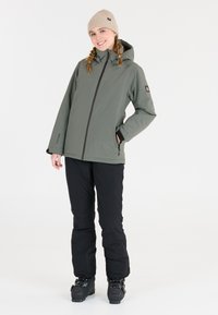 Grüne wasserdichte Jacke mit Kapuze, schwarze isolierte Hose und robuste schwarze Stiefel; verfügt über einen Reißverschluss und Seitentaschen.