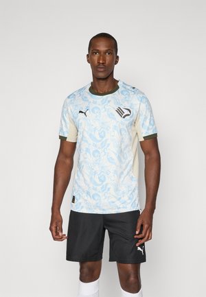 Mann trägt ein hellblaues gemustertes Sporttrikot mit Adler-Logo, schwarze Puma-Shorts und weiße Socken, steht vor einem einfarbigen Hintergrund.