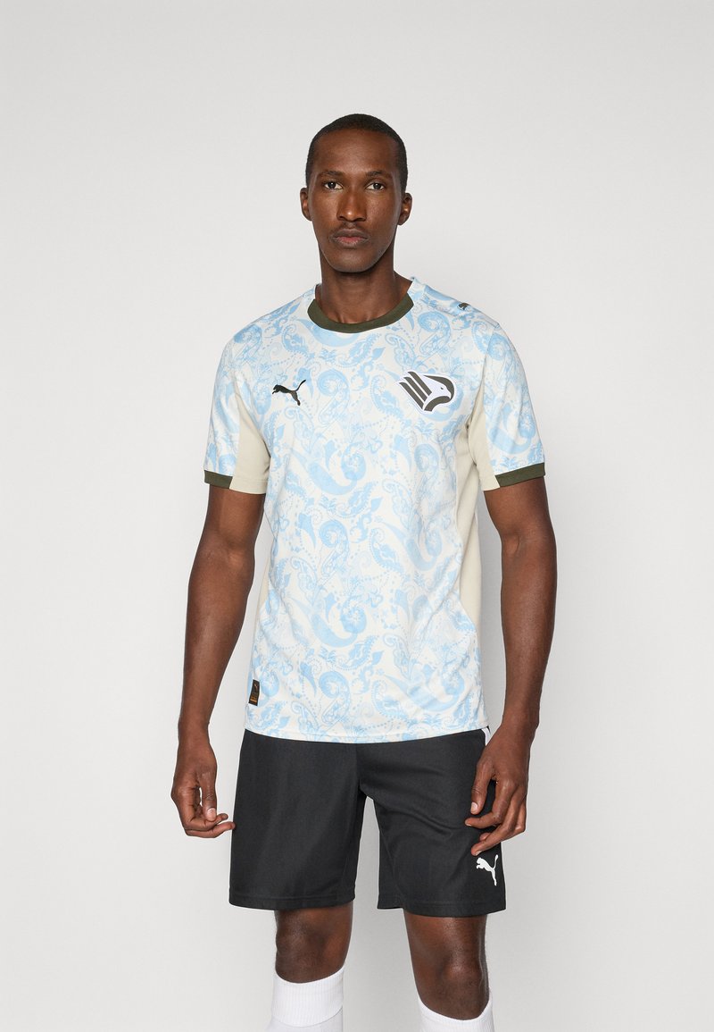 Uomo che indossa una maglia sportiva a fantasia blu chiaro con logo aquila, pantaloni corti neri Puma e calze bianche, in piedi contro uno sfondo semplice.